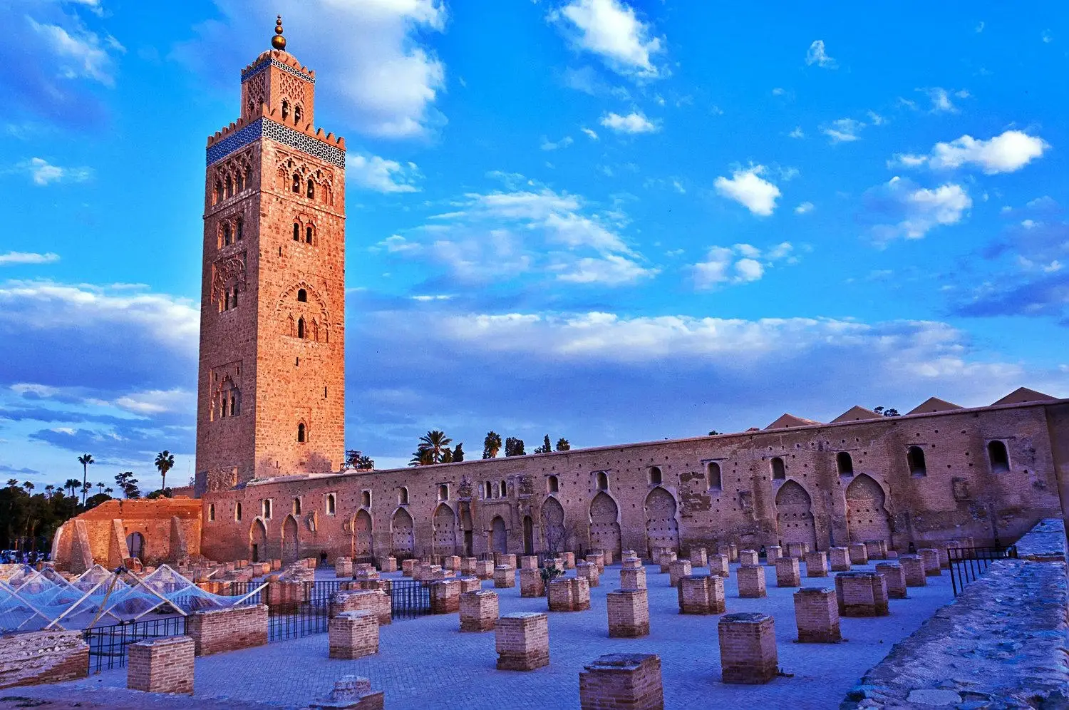 A bustling scene of Djemaa el‑Fna square at sunset in Marrakech, with dozens of white awnings covering market stalls, a crowd of people wandering lively alleys, and the silhouette of the Koutoubia Mosque minaret standing tall against a golden-pink sky.