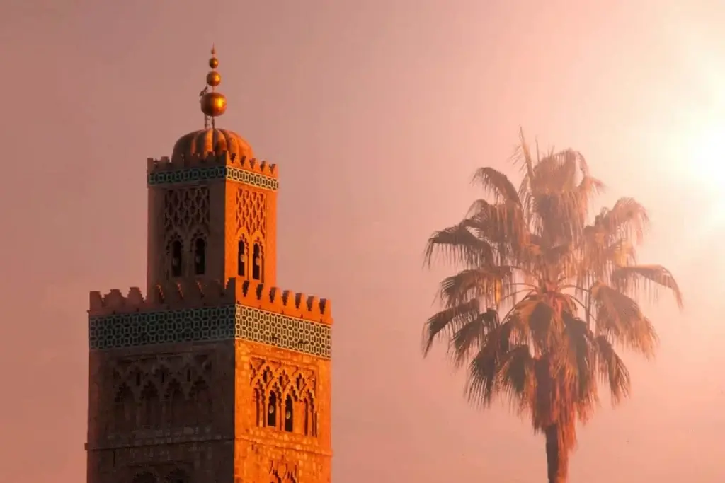 A bustling scene of Djemaa el‑Fna square at sunset in Marrakech, with dozens of white awnings covering market stalls, a crowd of people wandering lively alleys, and the silhouette of the Koutoubia Mosque minaret standing tall against a golden-pink sky.