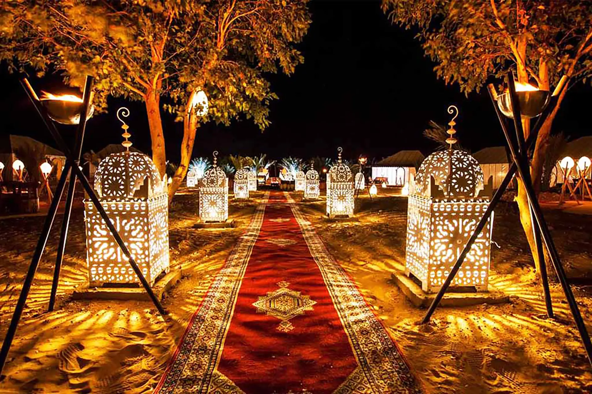 “Burrowed into the sand at the edge of Erg Chebbi, a desert camp with white‑canvas luxury tents, glowing lanterns, carpets, and low tables, framed by wind‑shaped golden dunes and a fiery sunset sky.”